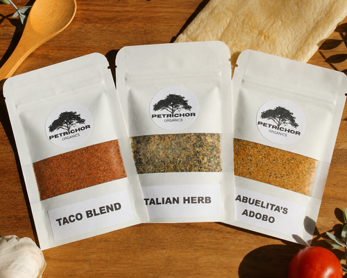 Three stand-up pouches of Petrichor Organics spices arranged on a wooden surface: Taco Blend, Italian Herb, and Abuelita's Adobo. The white bags have clear windows showing the spice blends inside, with a wooden spoon, garlic bulb, tomatoes, and olive leaves as styled props.